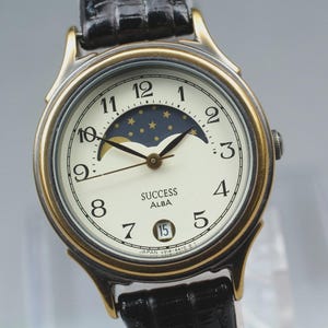 May include: An Alba Success wristwatch with a gold-tone case and a black leather strap. The cream-coloured dial features black Arabic numerals, a moon phase display with stars, a date window at the six o'clock position, and black hands.