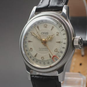May include: An Oris automatic watch with a silver case and a cream dial. The watch has gold hands and markers, a black leather strap, and a date display. The watch face has the text "ORIS 17 JEWELS AUTOMATIC SWISS MADE".