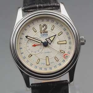 May include: A silver wristwatch with a cream face and black leather strap. The watch face displays black and blue numerals, a date indicator, and the text "ORIS AUTOMATIC". It has a second dial and a red-tipped hand.
