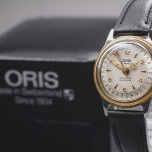 Może przedstawiać: Zegarek Oris w stylu vintage ze złotą ramką i czarnym skórzanym paskiem. Tarcza zegarka jest biała ze złotymi wskazówkami i cyframi. Zegarek jest prezentowany przed czarnym pudełkiem z logo i tekstem Oris.