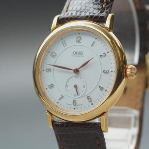 May include: A gold wristwatch with a white face and brown hands. The watch displays the brand name "ORIS" and "17 JEWELS". It has a small seconds dial and is attached to a brown leather strap.