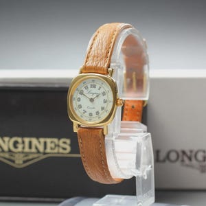 May include: A gold-toned Longines quartz watch with a light brown leather strap. The watch has a square face with a white dial and black numerals. The watch is displayed on a clear stand.