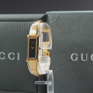Può includere: Un orologio Gucci color oro con quadrante nero rettangolare e lancette dorate. L'orologio è esposto su un supporto trasparente, con il logo Gucci visibile sullo sfondo. L'orologio ha un cinturino dorato e un design classico.