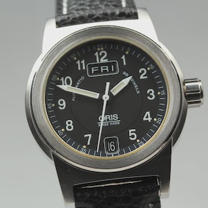 May include: A stainless steel wristwatch with a black dial, white numerals and hands. The watch shows the day as "FRI" and the date as "16". The watch has a black leather strap.