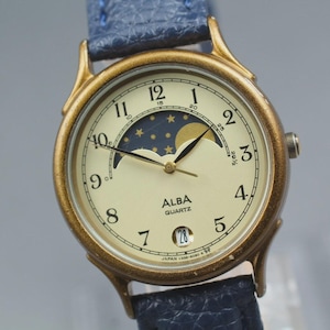 May include: An Alba quartz wristwatch with a gold-tone metal case and a textured blue leather strap. The cream-coloured dial features black Arabic numerals, a moon phase display with stars, a date window at the six o'clock position, and black hands.