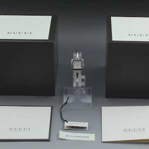 Può includere: Un orologio Gucci argentato con quadrante quadrato è esposto su un supporto trasparente. L'orologio è accompagnato da due scatole nere con coperchi bianchi, ciascuna con il logo Gucci. Sono inclusi documenti e un'etichetta.