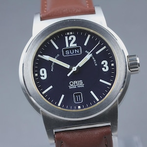 May include: A stainless steel wristwatch with a dark blue face, featuring white numerals and hands. The watch has a brown leather strap and the word "ORIS" is visible on the face. The watch also displays the day of the week.