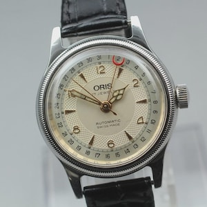 May include: A silver Oris wristwatch with a cream face and black leather strap. The watch face has gold hands and markers, with the text "ORIS 17 JEWELS AUTOMATIC SWISS MADE". The watch has a date display.