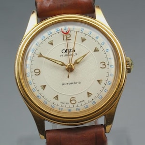 Op de afbeelding: Een vintage goudkleurig Oris polshorloge met een crèmekleurige wijzerplaat en bruine leren band. Het horloge heeft gouden wijzers en markeringen, een datumweergave en de tekst "ORIS 17 JEWELS AUTOMATIC". Een klassiek uurwerk.
