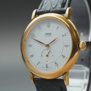 Puede incluir: Un reloj Oris dorado con esfera blanca y correa de cuero negro. El reloj tiene una subesfera en la parte inferior y agujas doradas. La esfera del reloj muestra el texto "ORIS 17 JEWELS".