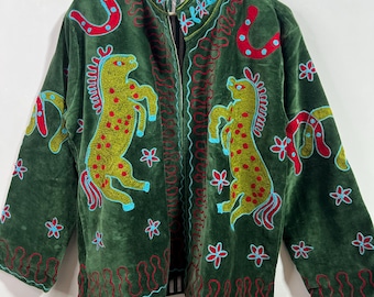 May include: A dark green jacket with colorful embroidery. The jacket features two embroidered horses, red horseshoes, and floral designs. The embroidery is in shades of red, blue, and yellow. The jacket is hanging on a wooden hanger.