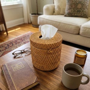 May include: A cylindrical woven rattan tissue box cover with a removable lid and a central opening, containing a white tissue. The natural light brown material features a tight, uniform weave pattern and a small decorative knot detail on the side.