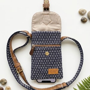 May include: A navy blue and white patterned crossbody phone bag with brown leather accents, a front zippered pocket, a snap button closure, and an adjustable strap. The interior lining is beige with a repeating logo print and a leather brand tag.