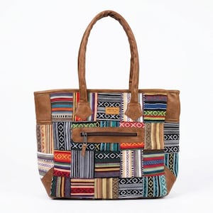 May include: A tote bag featuring a patchwork design of multi-colored woven fabric panels with geometric patterns. The bag includes brown faux leather handles, trim, and a zippered front pocket. A small rectangular label is centered near the top.