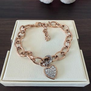 May include: A rose gold-toned chain link bracelet featuring a heart-shaped charm encrusted with clear crystals. The bracelet includes a lobster claw clasp and a small dangling charm near the closure, resting on a cream-coloured display box.