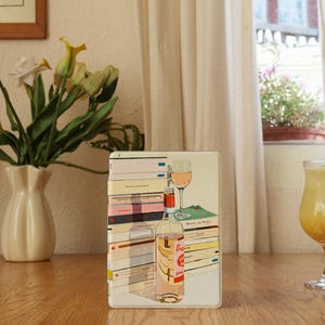 May include: A decorative metal box featuring a stack of books, a wine glass, and a bottle of rosé. The box is on a wooden table, with a vase of flowers and a glass of yellow beverage nearby. The background includes a window with a view.