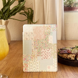 Puede incluir: Una funda para tableta digital con un diseño de patchwork en colores pastel. La funda presenta varios patrones florales, de cuadros y a rayas en rosa, verde y crema. La funda está de pie sobre una mesa de madera.