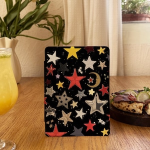 May include: Black tablet case decorated with stars in red, yellow, silver, and white. The case has a variety of star designs, including a crescent moon and an eye. The case is rectangular and stands upright on a wooden surface.