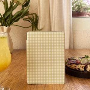 May include: A tablet case with a light green and white gingham pattern sits on a wooden table. A glass of yellow drink with a lime garnish and a pizza are also on the table. A vase of flowers and a window with curtains are in the background.