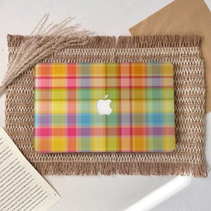Könnte beinhalten: Eine Laptop-Hülle mit einem farbenfrohen Karomuster in Gelb, Rosa, Grün und Blau. Das Apple-Logo ist sichtbar. Die Hülle liegt auf einer gewebten Matte mit Fransenrand.
