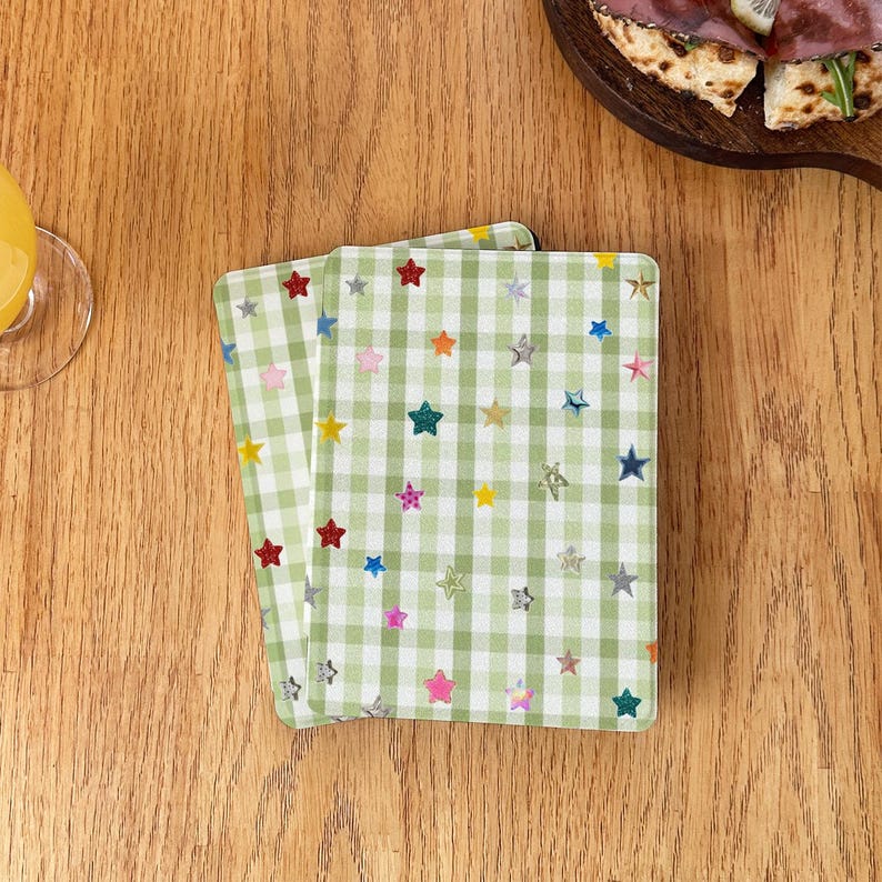 May include: Two rectangular coasters featuring a light green and white gingham check pattern. The surface of each coaster is decorated with a scattered print of small, multi-colored stars in shades of red, blue, yellow, pink, and silver.