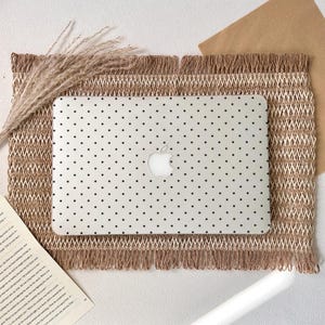 Könnte beinhalten: Ein weißer Laptop mit schwarzem Polka-Dot-Muster und Apple-Logo liegt auf einer beigen gewebten Tischmatte mit Fransen. Getrocknete Weizenähren und ein Buch sind ebenfalls im Bild zu sehen.