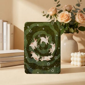 May include: A rectangular e-reader cover featuring a dark green swirling vortex pattern with five white rabbits and small white stars scattered across the surface. The design is centered on the front panel of the protective device case.