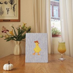 May include: A notebook featuring a blue and white striped design and a yellow duck wearing a brown cowboy hat. The notebook is on a wooden table. A white pumpkin and a vase of flowers are also on the table.