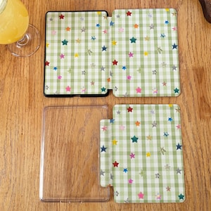 May include: A clear plastic protective case for an e-reader, featuring a light green and white gingham pattern decorated with small, colorful, multi-pointed stars. The set includes a front cover, a back cover, and a separate clear frame piece.