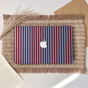 Puede incluir: Funda para portátil con un patrón de rayas verticales en burdeos y azul claro. El logotipo de Apple está centrado en la funda. La funda está sobre una estera tejida con borde de flecos, con trigo seco y un libro en el fondo.