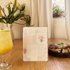 May include: A pastel-colored e-reader case with a patchwork design featuring pink floral accents and a tulip illustration. The case is standing upright on a wooden table, with a glass of yellow liquid and a pizza in the background.