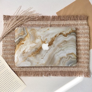 Peut inclure: Une coque rigide pour ordinateur portable arborant un motif de marbre blanc et doré avec des veines tourbillonnantes et une découpe centrale pour le logo Apple. La surface présente une finition brillante aux motifs organiques complexes.