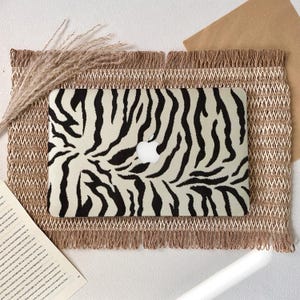 Puede incluir: Un ordenador portátil con una funda con estampado de cebra, con rayas negras sobre un fondo crema. El portátil está colocado sobre un mantel individual beige tejido con un borde con flecos. También hay una ramita de hierba seca y un libro.