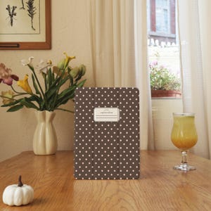 May include: A brown tablet case with a polka dot pattern and a rectangular label. The case is on a wooden table, next to a vase of flowers, a glass of juice, and a small white pumpkin.