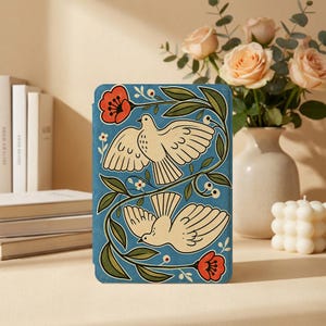 May include: A rectangular tablet case featuring a blue background with two cream-colored birds in flight, surrounded by green leafy vines and red flowers. The design is symmetrical, with one bird facing upward and the other facing downward.