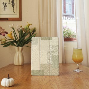 Könnte beinhalten: Eine iPad-Hülle mit einem Patchwork-Design in Creme-, Beige- und Salbeigrün-Tönen mit floralen und geometrischen Mustern. Die Hülle liegt auf einem Holztisch, zusammen mit einer Blumenvase, einem Glas Saft und einem kleinen weißen Kürbis.