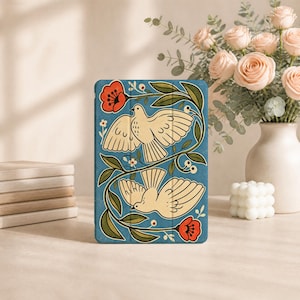 Può includere: Una custodia rettangolare per tablet con sfondo blu, decorata con due uccelli color crema in volo, circondati da tralci di foglie verdi e fiori rossi. Il design è simmetrico, con un uccello posizionato sopra l'altro, entrambi raffigurati con ali e piume stilizzate.