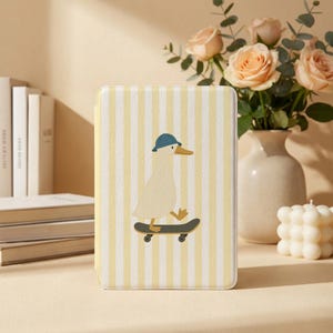 May include: A rectangular e-reader case featuring a light yellow and white vertical striped pattern. Centered on the front is a graphic illustration of a white duck wearing a blue cap while riding a black skateboard on a light background.
