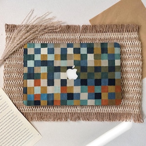 May include: A laptop case with a colorful checkered pattern in shades of blue, green, orange, and tan. The Apple logo is visible. The case rests on a woven mat with a fringed edge, and dried grass.