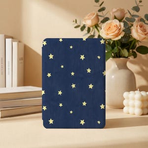 Pode incluir: Um objeto retangular azul marinho com um padrão de estrelas amarelas. O objeto está em pé sobre uma superfície bege. Ao fundo, há livros, um vaso de rosas e uma vela branca.