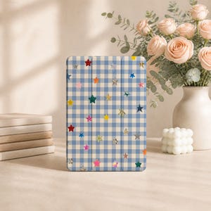 May include: A rectangular tablet case featuring a blue and white gingham check pattern decorated with scattered, multi-coloured glittery star shapes in various sizes. The case has a tri-fold design with visible vertical creases across the front surface.