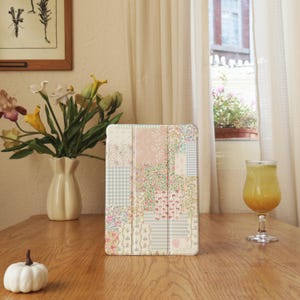 Peut inclure: Une housse de tablette à motifs floraux avec un design patchwork aux couleurs pastel, notamment rose, vert et blanc. La housse est posée sur une table en bois, à côté d'un vase de fleurs, d'une petite citrouille et d'un verre de jus.