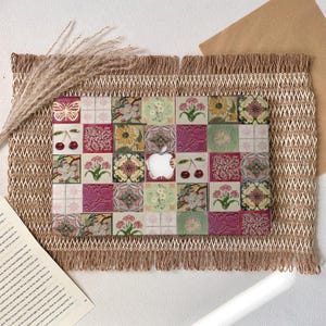 May include: A decorative laptop case featuring a patchwork design of floral and fruit tiles in shades of pink, green, and yellow. The Apple logo is visible in the center. The case is displayed on a textured, fringed mat with dried grass.