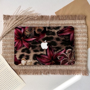 Puede incluir: Funda para portátil con estampado de leopardo y detalles florales en rojo oscuro. La funda tiene un logotipo blanco de Apple. Se encuentra sobre un mantel individual tejido con bordes con flecos.