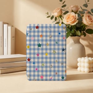 Puede incluir: Una funda rectangular para lector electrónico con un patrón de cuadros vichy en azul claro y blanco. La superficie está decorada con numerosas estrellas pequeñas y multicolores en varios tonos, incluyendo rojo, amarillo, verde, rosa y plata, dispersas por la cuadrícula.
