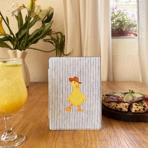Könnte beinhalten: Ein rechteckiges Notizbuch mit blau-weißem Streifenmuster. Auf dem Cover ist eine gelbe Ente mit braunem Hut abgebildet. Das Notizbuch liegt auf einem Holztisch. Ein Glas gelbes Getränk und eine Pizza sind im Hintergrund.