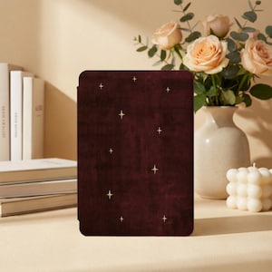 May include: A rectangular e-reader case in a deep burgundy velvet texture, featuring a scattered pattern of small, four-pointed gold stars across the front surface. The case has a smooth, solid-colored finish with a soft, tactile appearance.