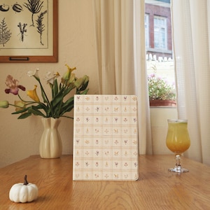 Può includere: Un quaderno color crema con un motivo floreale è appoggiato su un tavolo di legno. Il quaderno è circondato da un vaso di fiori, una piccola zucca bianca e un bicchiere di succo d'arancia. Una stampa botanica incorniciata è appesa al muro.