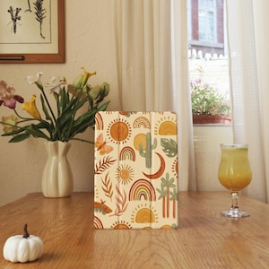 May include: A beige tablet case featuring a repeating pattern of suns, rainbows, cacti, butterflies, and leaves in shades of orange, brown, and green. The case is on a wooden table with a vase of flowers and a glass of juice.