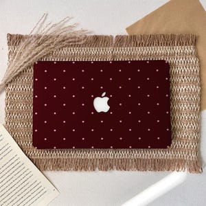 Puede incluir: Una funda para portátil de color burdeos con el logotipo blanco de Apple y pequeños lunares rosas claros espaciados uniformemente. La funda está sobre un mantel individual tejido y con flecos.